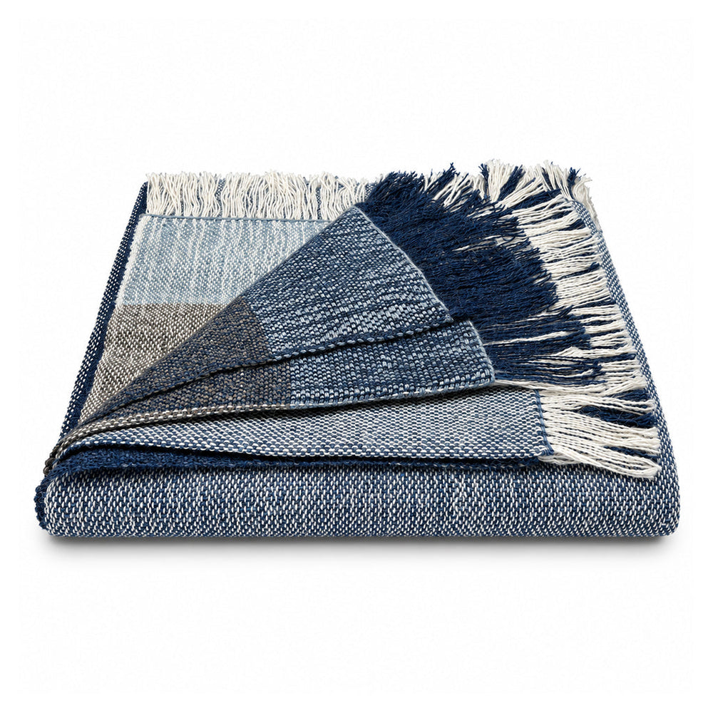 A folded woven throw blanket with a plaid pattern in shades of navy, blue, gray, and white, featuring cream-colored fringe on one end, set against a plain white background.