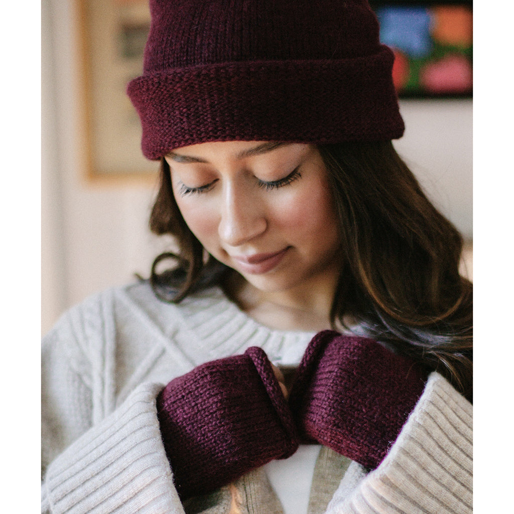 A woman wearing a dark burgundy knit beanie and matching knit fingerless mittens, paired with a cream-colored ribbed sweater, looking down with a soft expression.