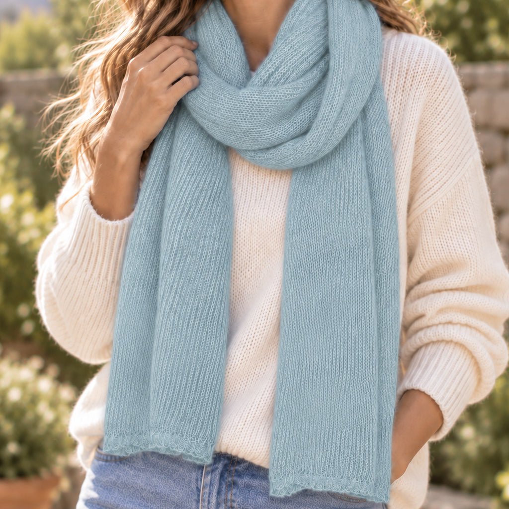 A person wearing a light blue knit scarf over a cream-colored knit sweater, photographed outdoors with blurred greenery in the background.