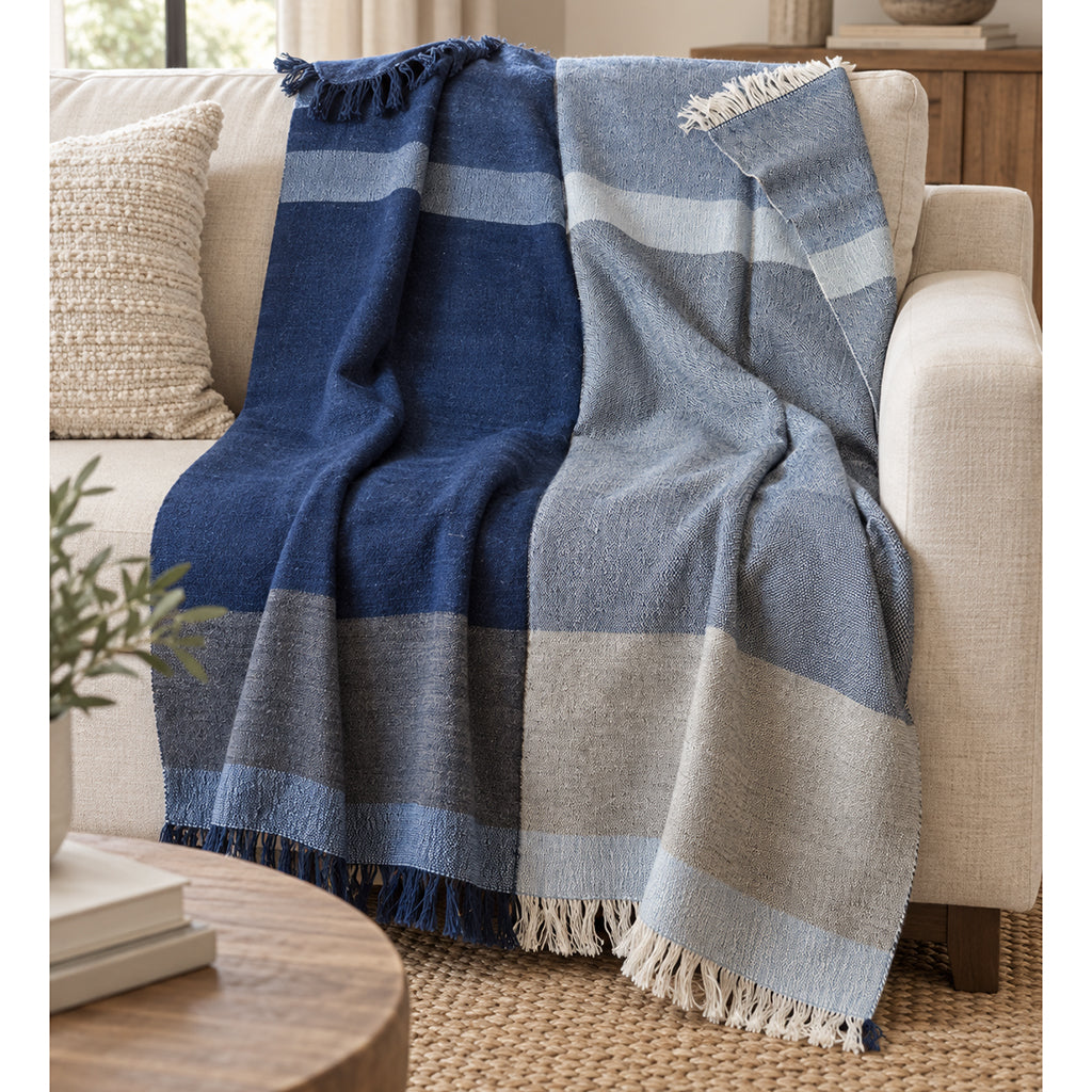 A woven throw blanket with large blue, light blue, and beige stripes and fringed edges, draped over a cream-colored armchair in a cozy living room setting.