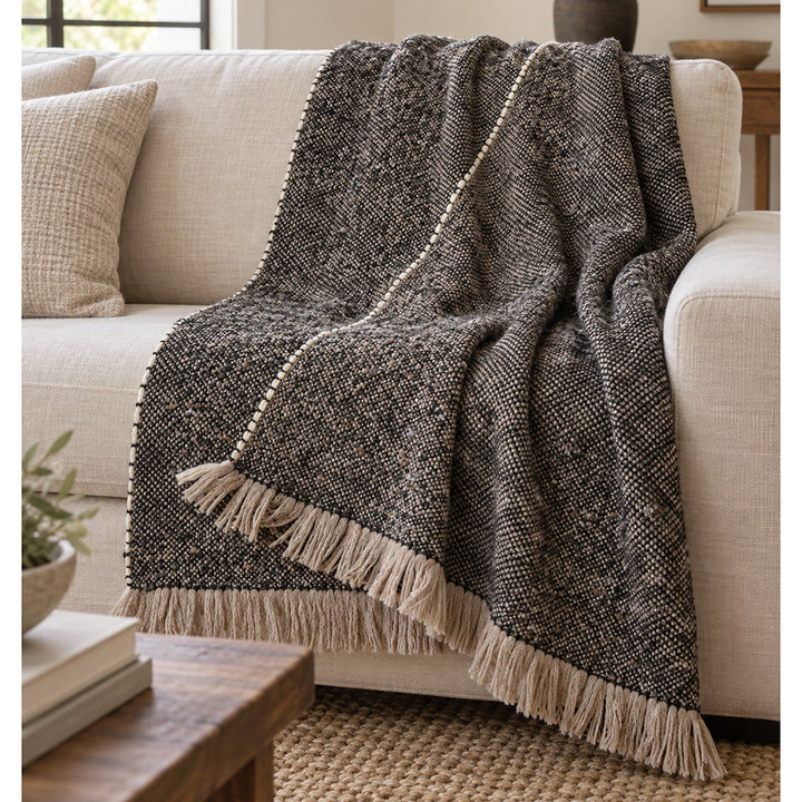 A dark charcoal and beige woven alpaca blend throw blanket with fringe trim draped over a light beige sofa in a cozy living room setting.