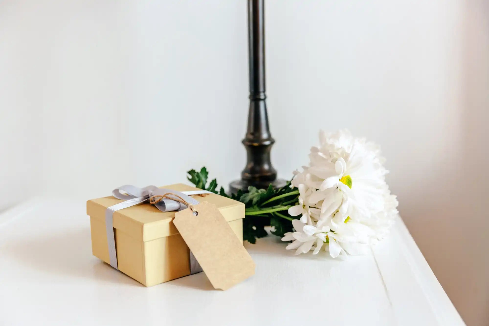 Gift box with a tag attached, placed next to white flowers and a candlestick.