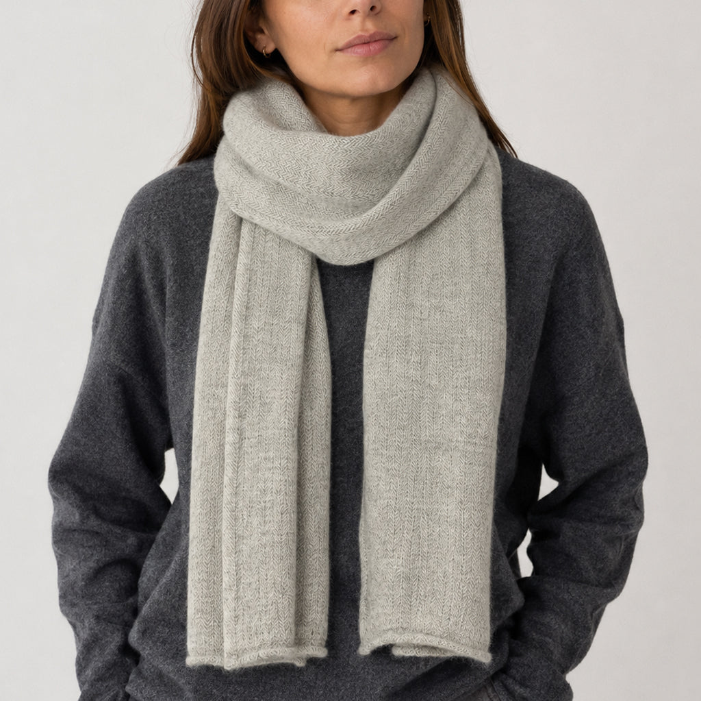 A woman wearing a light gray knit scarf with herringbone pattern wrapped around her neck over a dark gray sweater.