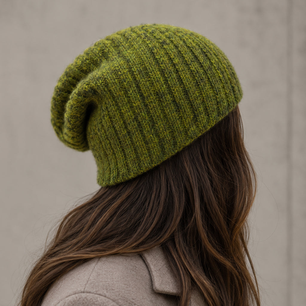A person shown from the back wearing a slouchy olive green ribbed knit beanie hat, with long brown hair and a beige coat against a neutral gray wall.