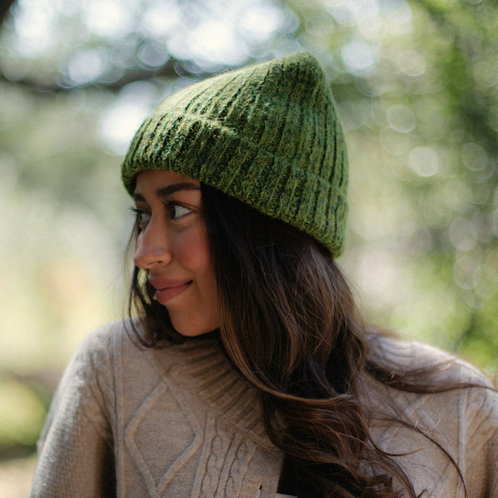 A woman wearing a textured olive green knit beanie hat and a beige cable-knit sweater, photographed outdoors with a soft, blurred natural background.
