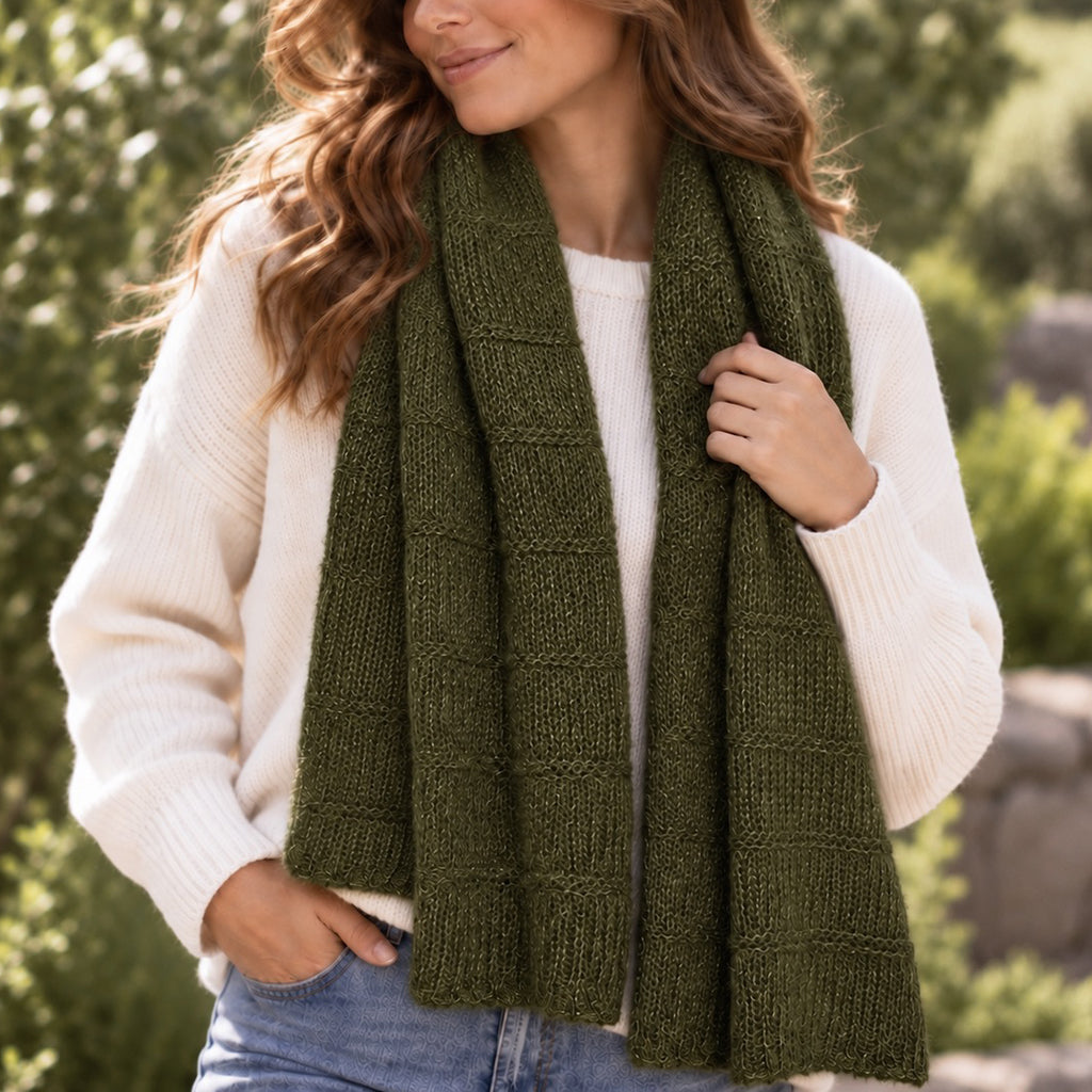 A woman outdoors wearing a chunky olive green knit scarf draped over a cream ribbed sweater and blue jeans.