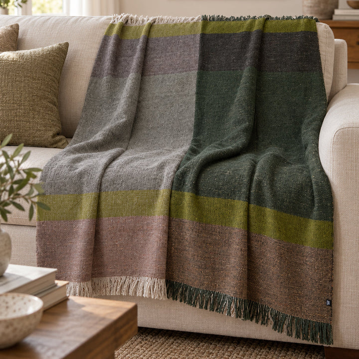 A woven throw blanket with a large check pattern in gray, olive green, brown, and muted pink, draped over a light beige sofa in a cozy living room setting.