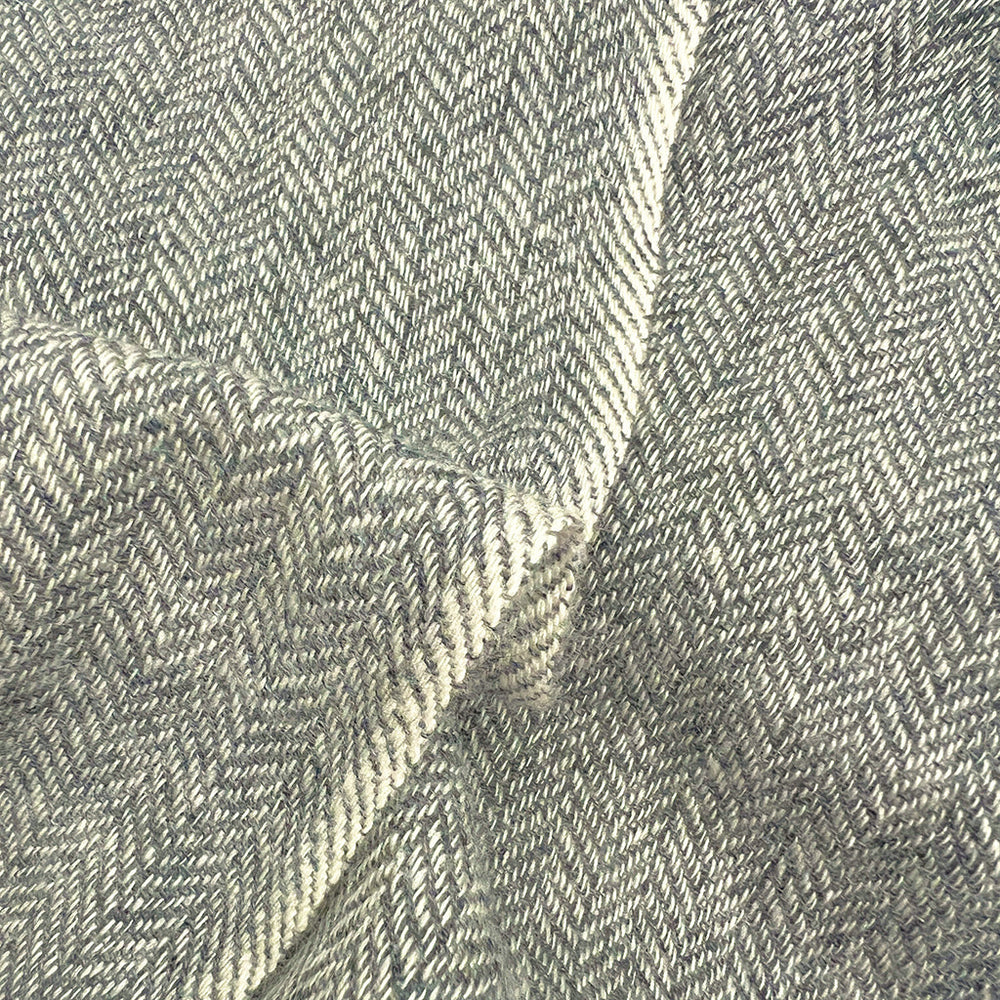 A close-up of a woven fabric with a herringbone pattern in light gray and off-white, showing the texture and folds of the material.