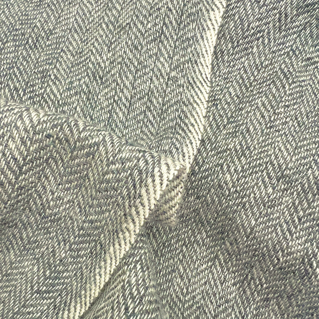 A close-up of a woven fabric with a herringbone pattern in light gray and off-white, showing the texture and folds of the material.