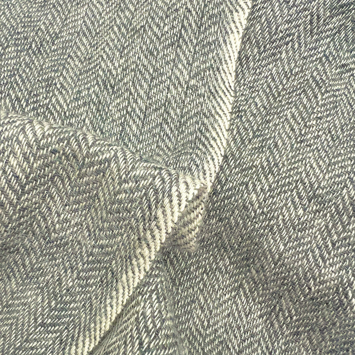 A close-up of a woven fabric with a herringbone pattern in light gray and off-white, showing the texture and folds of the material.